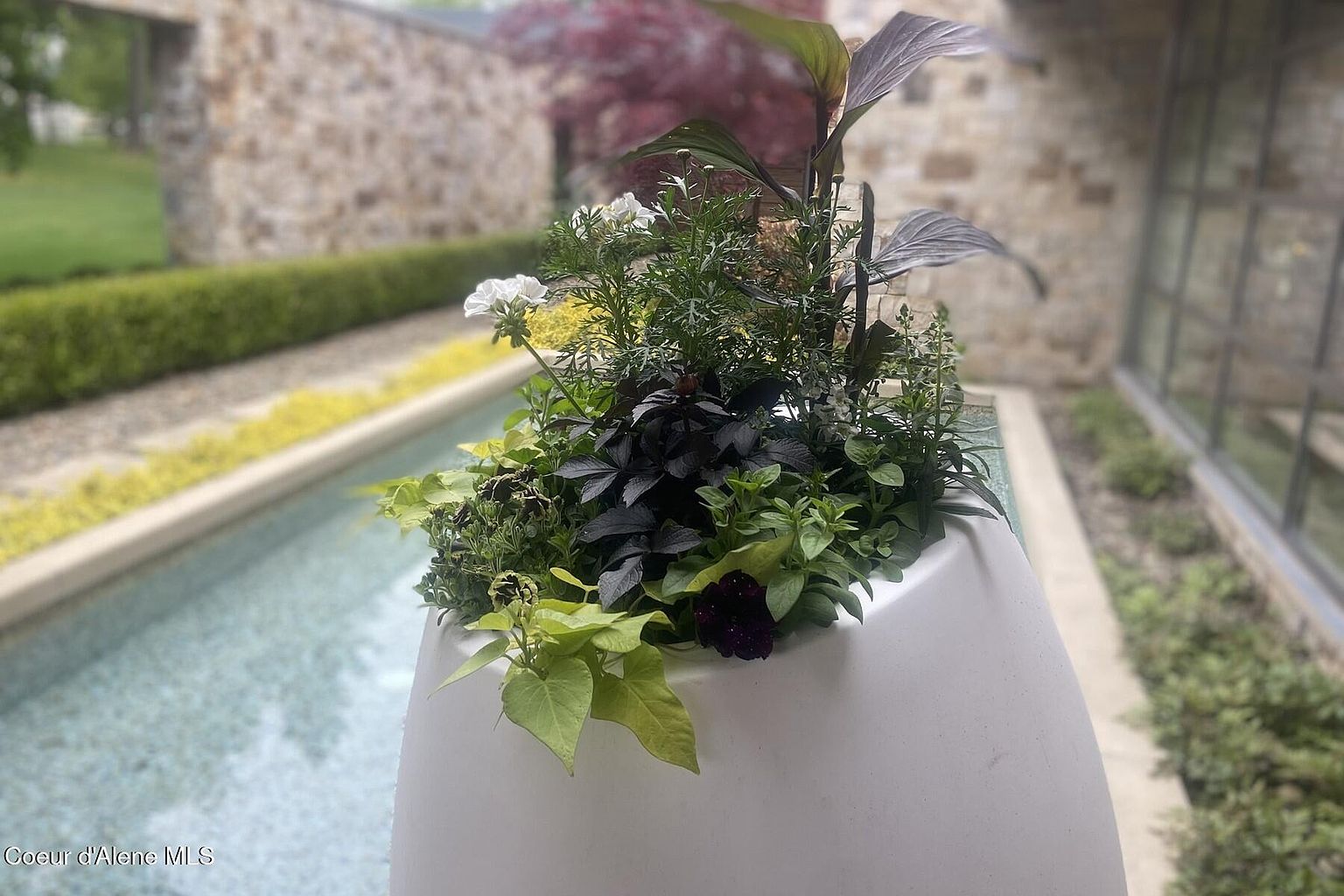 This image showcases a beautifully landscaped garden area, featuring a modern white planter filled with a variety of plants and flowers. A narrow water feature adds a touch of serenity, while a stone wall and manicured hedges provide a structured backdrop. The overall impression is one of tranquility and sophisticated outdoor living.