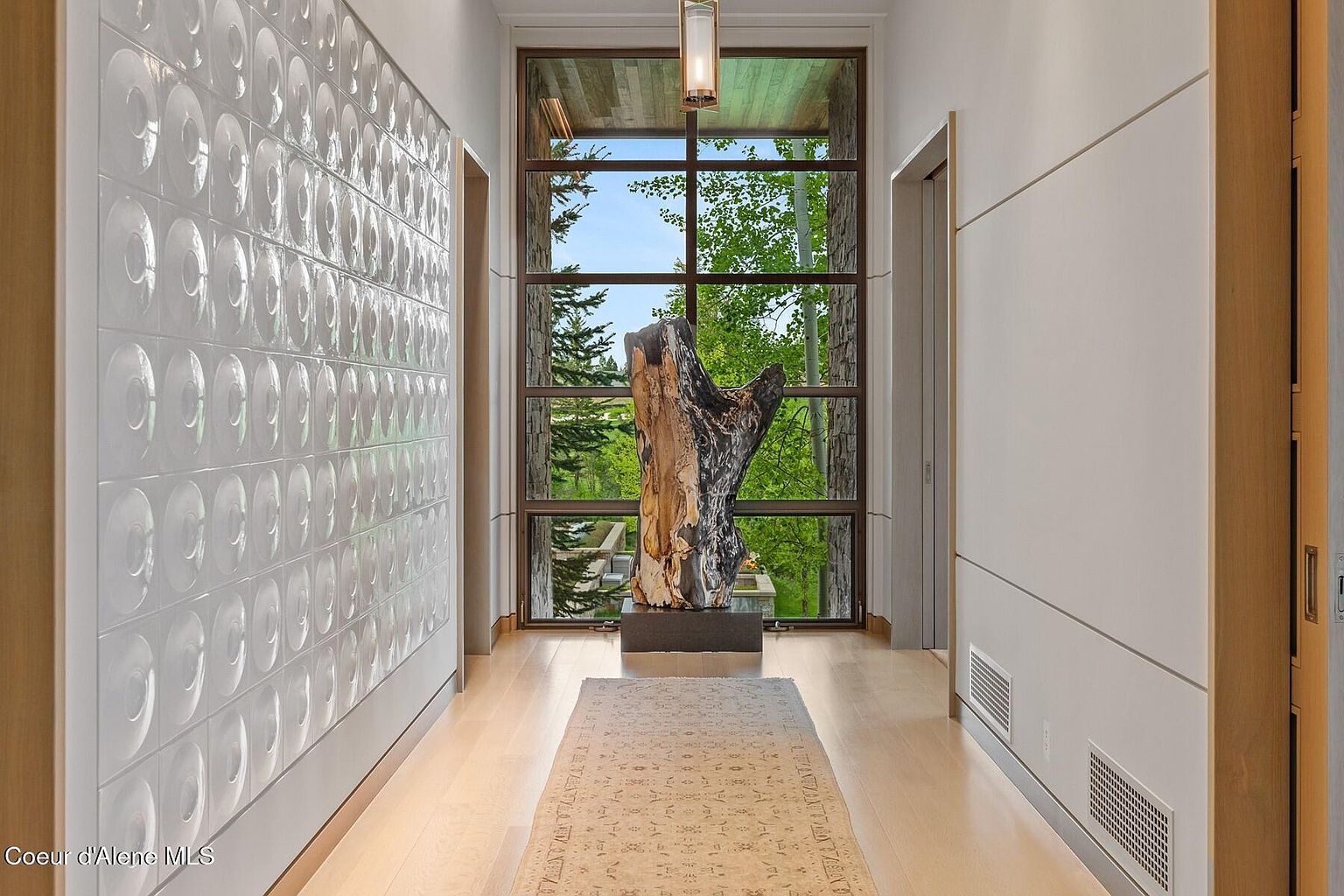 This interior shot showcases a modern hallway with unique architectural details. A textured white wall contrasts with the smooth, light-colored wood flooring and a large window at the end of the hall, framing a view of lush greenery and a striking tree sculpture. The hallway is well-lit, creating a bright and inviting space.