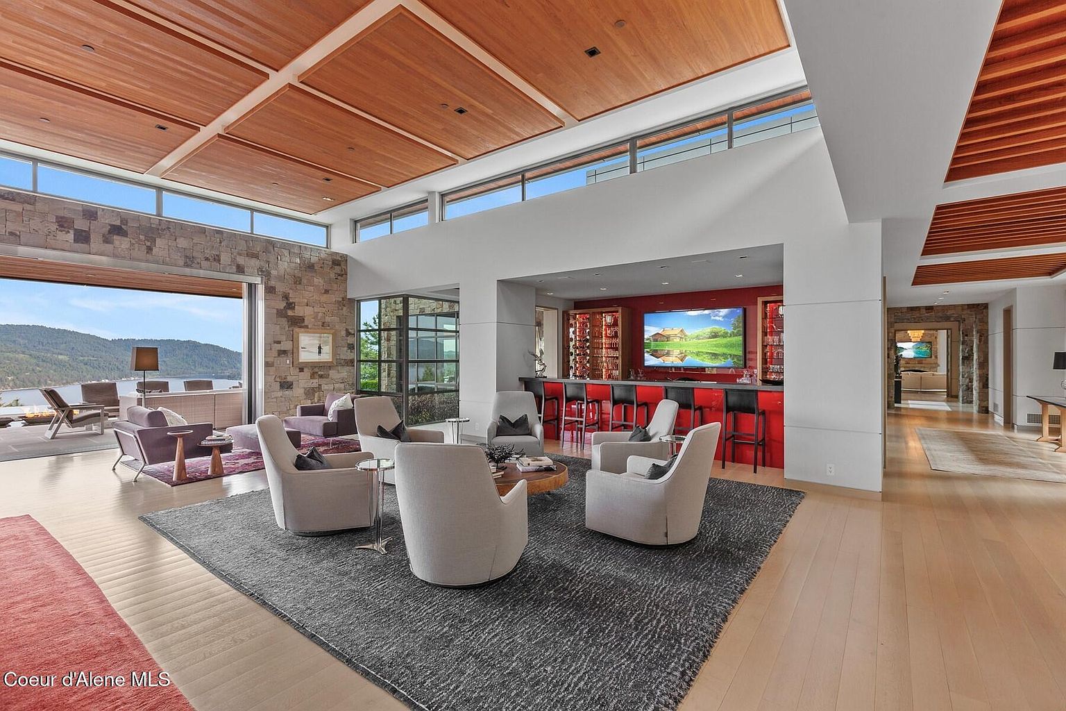 This expansive living room features a modern design with a wood-paneled ceiling and a stone accent wall. A seating area with multiple armchairs is arranged around a central coffee table, and a bar area with red accents is visible in the background. Large windows offer a view of the lake, creating a luxurious and inviting atmosphere.