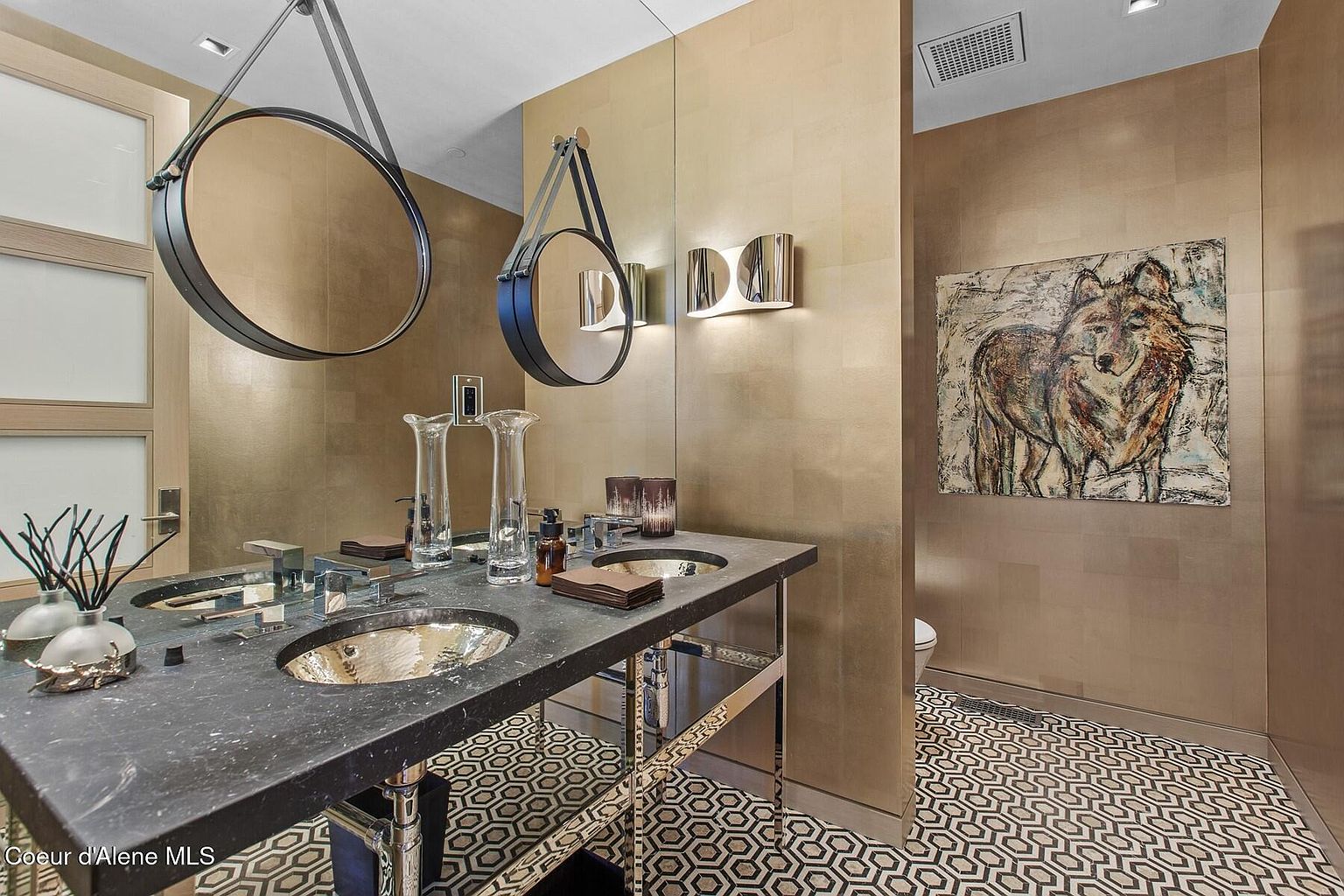 This is a stylish guest bathroom featuring a double vanity with a black countertop and unique round mirrors suspended from the ceiling. The walls are covered in a gold-toned tile, and the floor has a geometric pattern. An abstract painting of a wolf adds a touch of character to the space, creating a luxurious and modern feel.