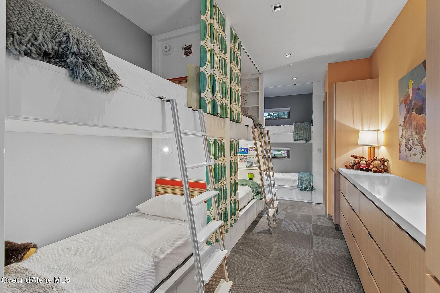 This is an interior shot of a bedroom featuring built-in bunk beds. The room has a modern design with white and light wood finishes, complemented by patterned curtains for privacy. The space is well-lit and appears to be designed for children or guests, offering a functional and stylish sleeping arrangement.