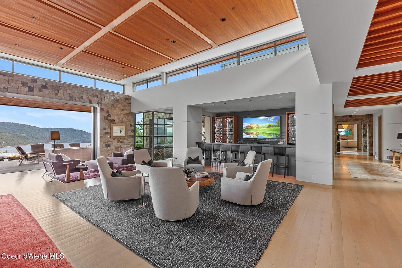 This is a spacious living room featuring a modern design with a high ceiling and wooden panels. The room includes a seating area with several armchairs arranged around a coffee table on a gray rug, and a bar area with stools and a large screen. Large windows offer a view of the lake, and the open layout connects to a hallway.