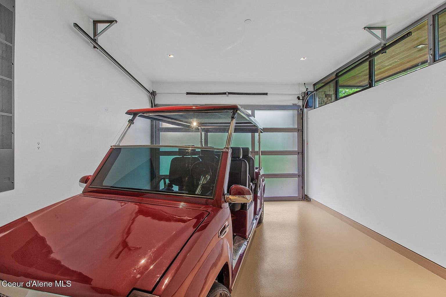 The image showcases a well-lit garage featuring a red golf cart parked inside. The garage has a modern aesthetic with a glass panel garage door and clean, white walls. The floor is a light tan color, and there are windows along the upper right side of the wall, adding natural light to the space.