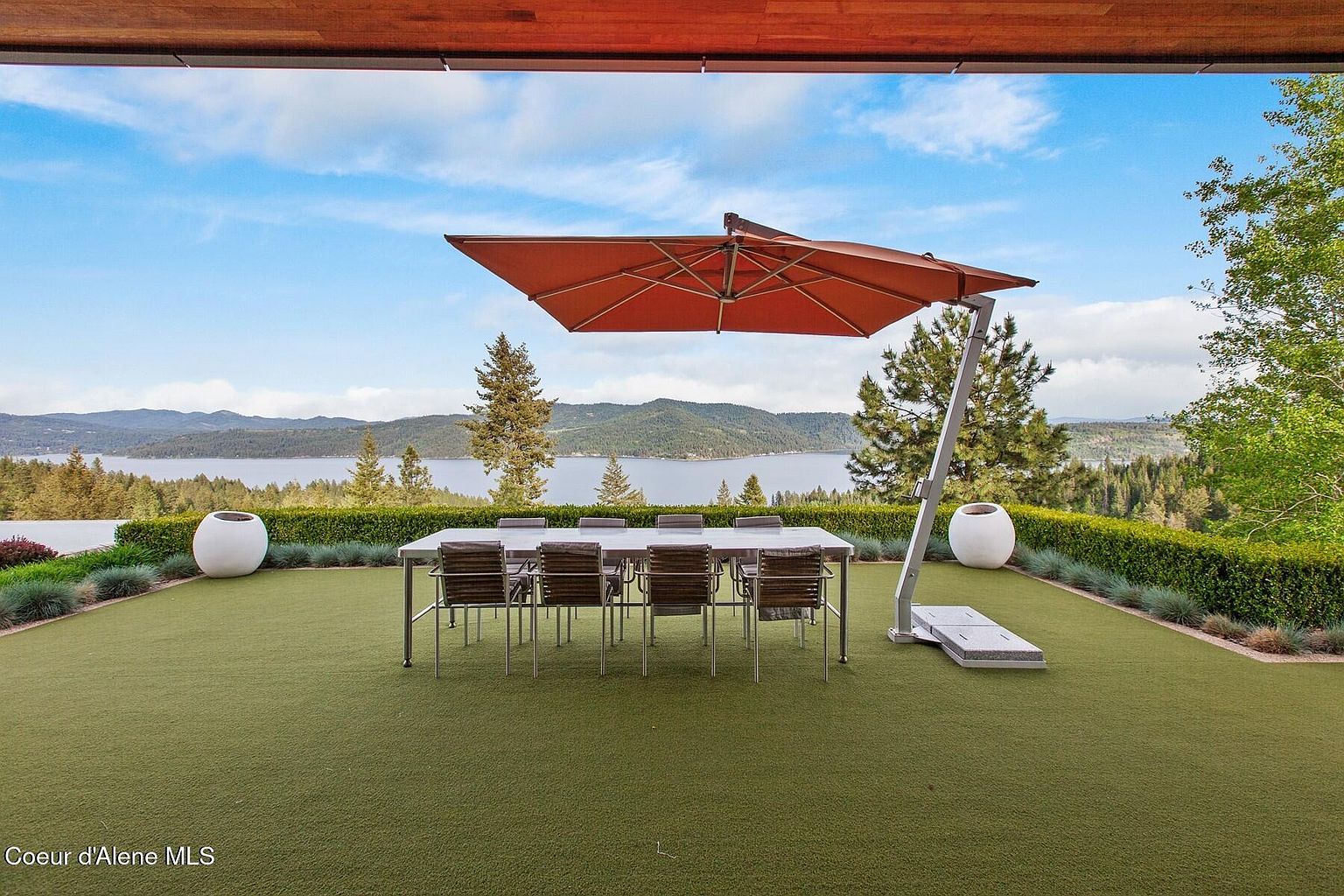 This image showcases a luxurious outdoor patio or deck area with a stunning view of a lake and mountains. A large dining table with chairs is centered under a cantilevered umbrella, flanked by decorative planters. The manicured lawn and surrounding greenery create a serene and inviting atmosphere, perfect for outdoor entertaining.