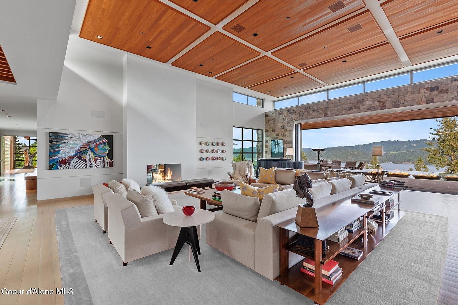 This is a spacious living room featuring a modern design with a wood-paneled ceiling and large windows offering a scenic view of a lake and mountains. The room is furnished with comfortable sofas, a fireplace, and a long console table behind the sofa, creating a luxurious and inviting atmosphere. An artwork of a Native American chief is displayed on the wall.