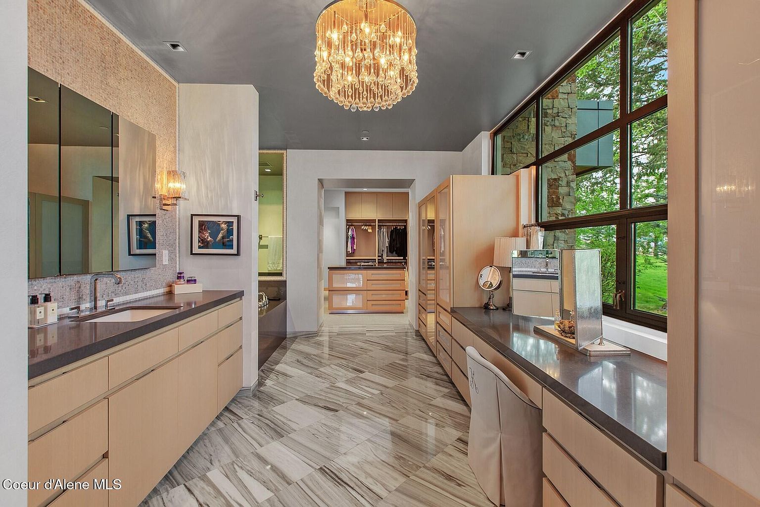 This is a luxurious primary bathroom featuring a double vanity with a dark countertop and light wood cabinetry. A large window provides natural light and a view of the outdoors. The bathroom also includes a walk-in closet and a chandelier, adding to the upscale feel of the space. The flooring is a light marble tile with dark veining.