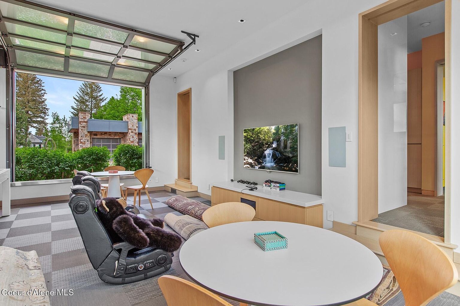 This interior shot showcases a modern living room with a unique indoor-outdoor feel, thanks to a large glass garage door that opens to the exterior. The room features a neutral color palette with a gray accent wall, a flat-screen TV, and comfortable seating arrangements, including a gaming chair and round tables, creating a versatile space for relaxation and entertainment. The checkered floor adds a touch of visual interest.