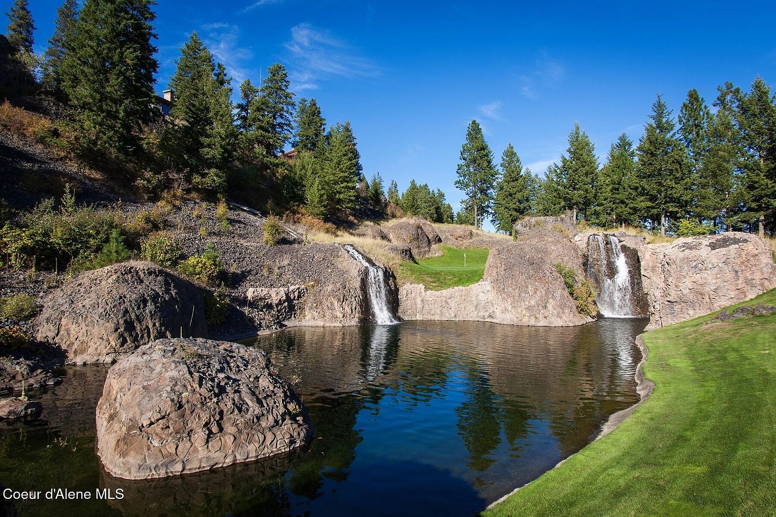 This picturesque outdoor scene showcases a beautifully landscaped yard featuring a pond with waterfalls cascading from rocky cliffs. Lush green grass surrounds the pond, and mature trees provide a scenic backdrop. The image evokes a sense of tranquility and natural beauty, highlighting the property's unique outdoor features.