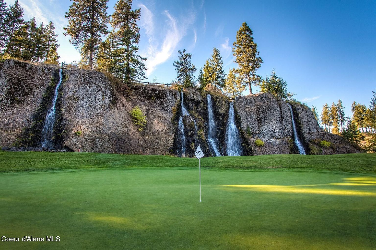 This image showcases a picturesque golf course amenity, featuring a lush green putting surface with a flag in the foreground. Behind the green, a rocky cliff face is adorned with multiple cascading waterfalls and mature trees, creating a stunning natural backdrop. The clear blue sky and wispy clouds enhance the serene and luxurious atmosphere of this unique golfing environment.