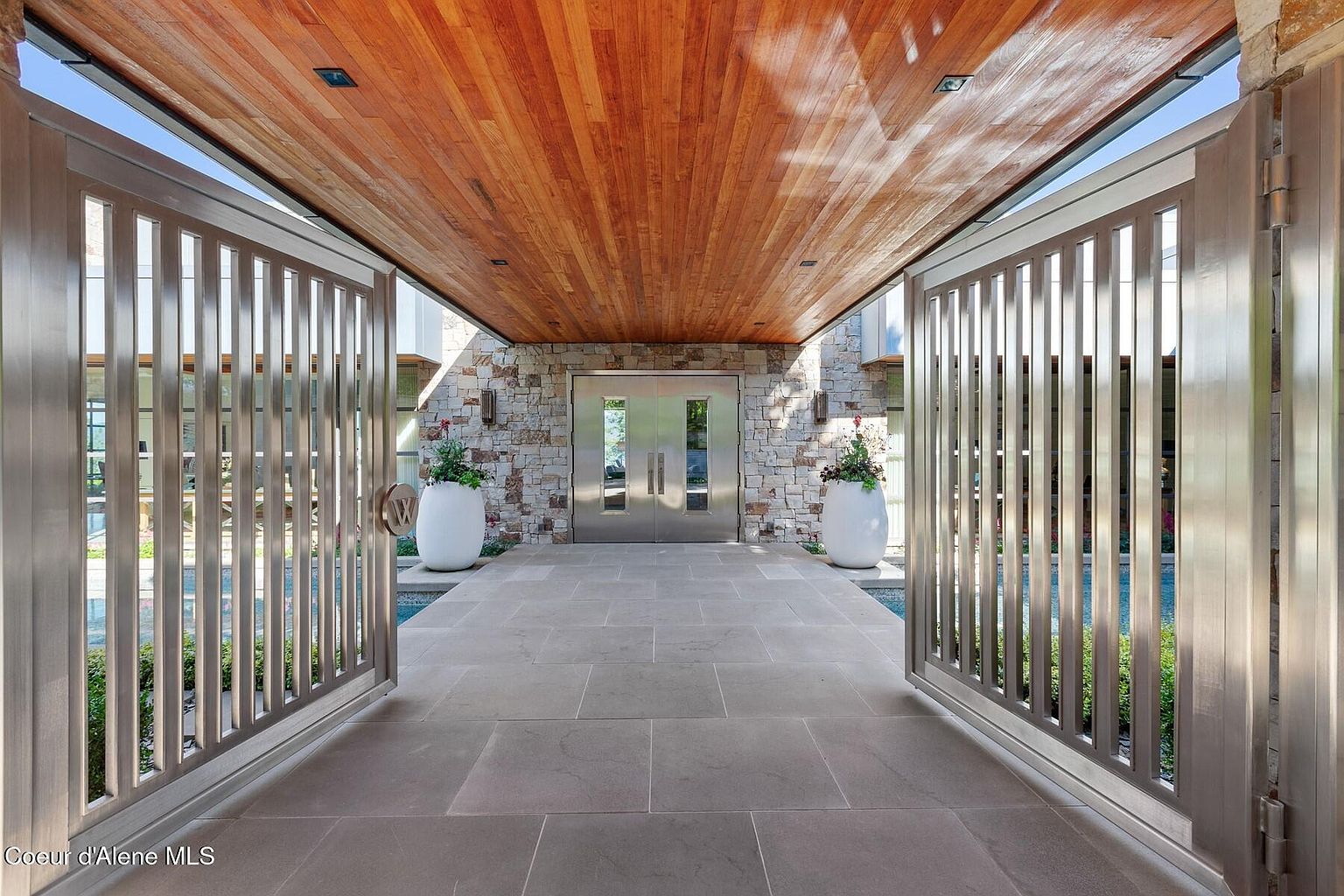 This image showcases a grand entryway to a luxurious home. The entrance features a striking combination of materials, including a warm wood ceiling, stone walls, and modern metal gates. Large planters flank the double doors, adding a touch of greenery and elegance to the sophisticated design.