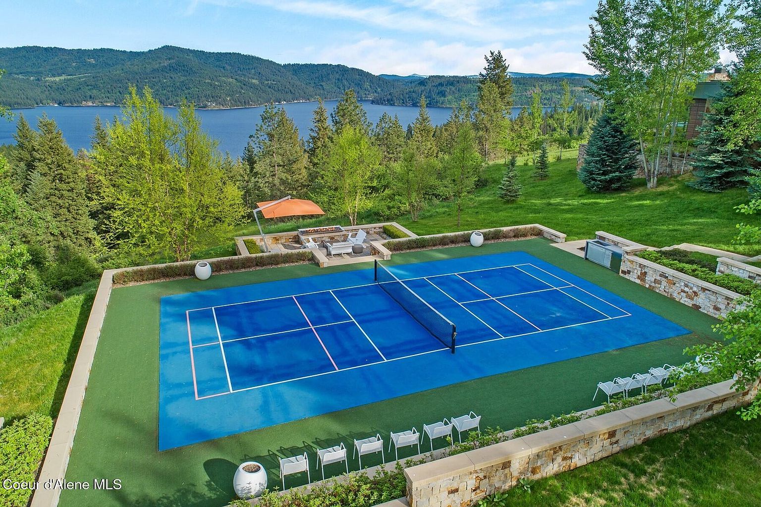 This aerial shot showcases a meticulously maintained yard featuring a vibrant blue tennis court with white lines, surrounded by lush green grass and manicured landscaping. A stone patio with outdoor seating and an orange umbrella provides a relaxing space, while the backdrop of a serene lake and forested hills adds to the property's appeal. The overall impression is one of luxury and recreational opportunities.