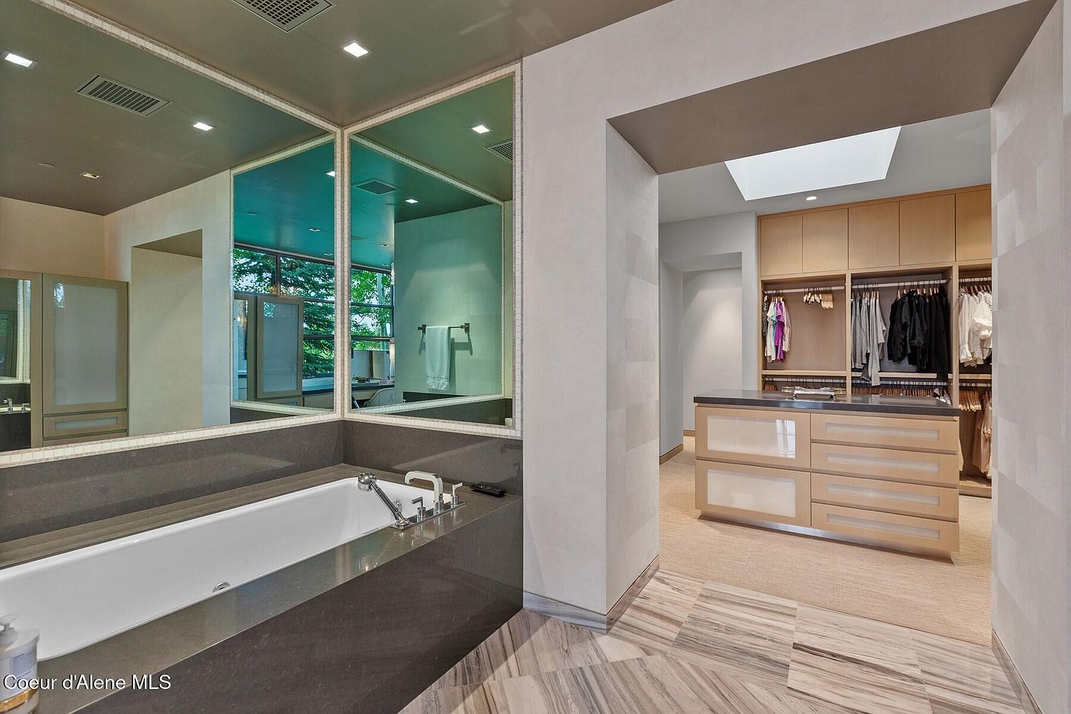 This image showcases a luxurious primary bathroom with a modern aesthetic. A large soaking tub is framed by a dark countertop and a mirrored wall, reflecting natural light. The bathroom seamlessly transitions into a walk-in closet, featuring custom cabinetry and a central island, creating a spa-like and functional space.