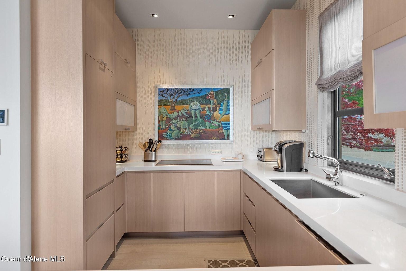 This is a well-lit, modern kitchen with light wood cabinetry and white countertops. The kitchen features a stainless steel sink, a window with a view of greenery, and an abstract art piece on the wall. The overall impression is clean, contemporary, and functional.