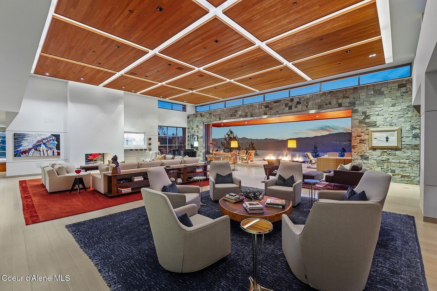 This is a spacious and luxurious living room featuring a modern design with a wood-paneled ceiling and a stone accent wall with a large sliding glass door that opens to an outdoor patio. The room is furnished with multiple seating areas, including a circular arrangement of armchairs on a dark blue rug and a sectional sofa on a red rug, creating a comfortable and inviting atmosphere. The open floor plan and natural light enhance the sense of grandeur and sophistication.