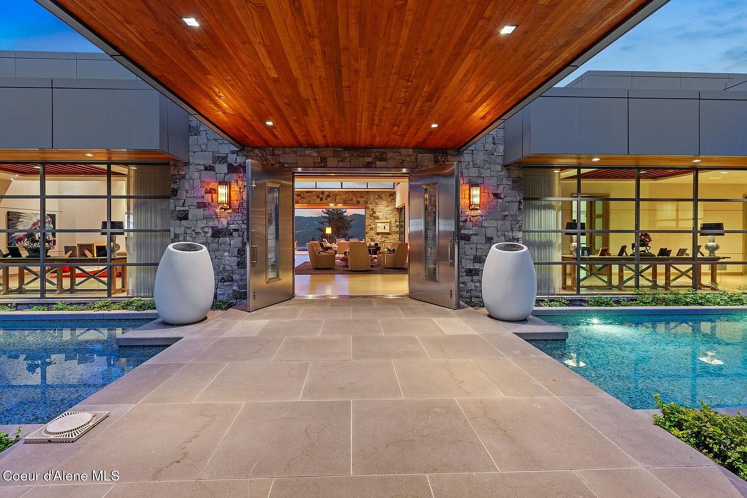 This is a striking entryway to a modern home, featuring a stone facade, a warm wood ceiling, and large stainless steel double doors that are open to reveal a glimpse of the interior living space. Two large, white, egg-shaped planters flank the entrance, which is bordered by a reflecting pool on either side, adding to the luxurious and contemporary feel of the property. The perspective is a wide shot, emphasizing the grand scale and inviting nature of the entrance.