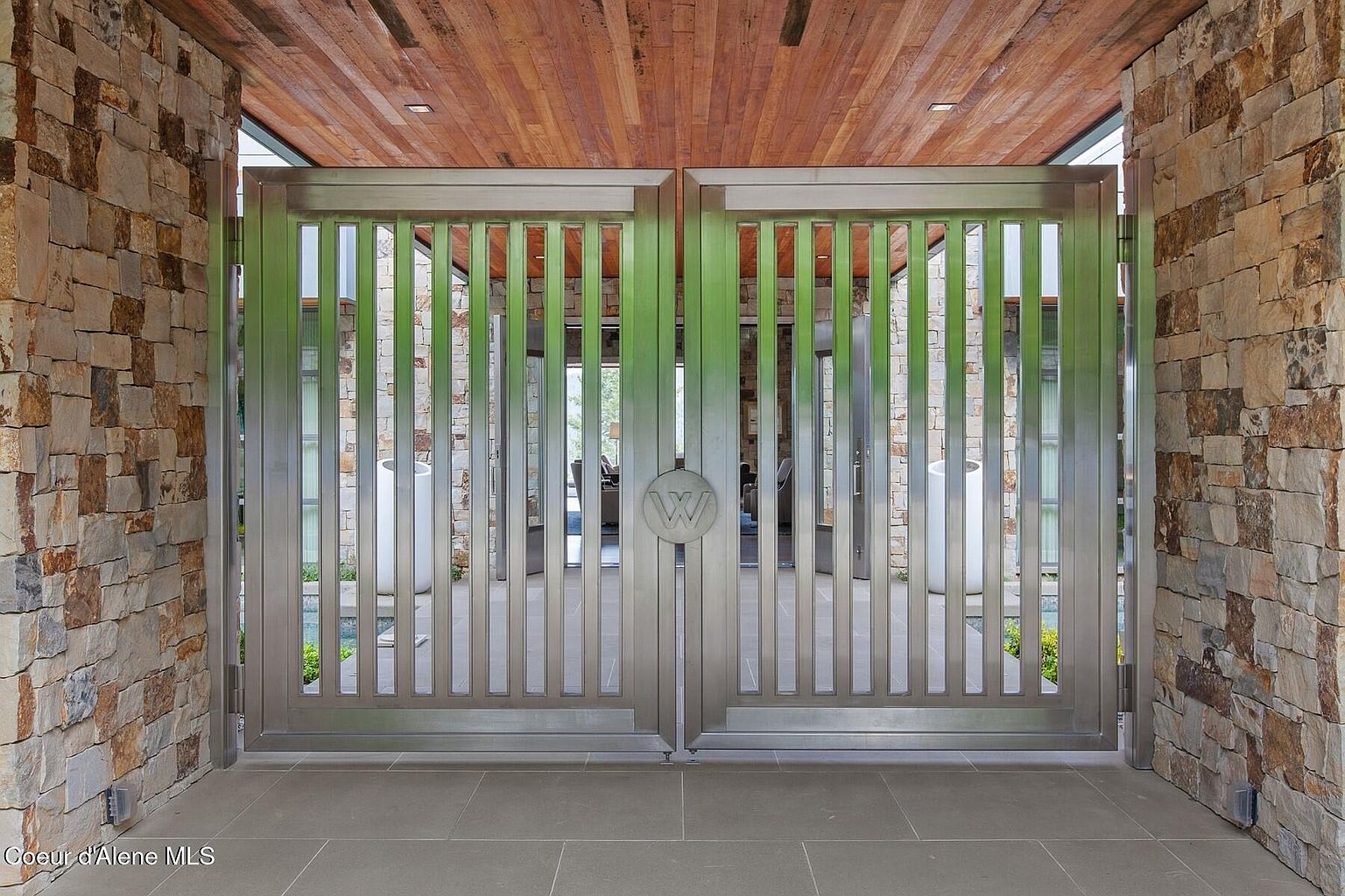 The image showcases a grand entryway featuring a pair of modern metal gates adorned with a circular emblem. The gates are flanked by stone walls, creating a rustic yet contemporary aesthetic. Above, a wooden ceiling adds warmth to the space, while the gray stone flooring provides a sleek foundation. The perspective is from the front, emphasizing the symmetry and scale of the entrance.