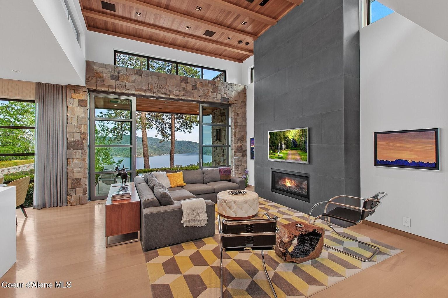 This is a modern living room with a large gray sectional sofa, a stone accent wall with large windows overlooking a lake, and a gray tiled fireplace with a mounted television. The room features a geometric patterned rug, a unique wooden side table, and a modern chair, creating a stylish and comfortable living space with stunning views. The perspective is from the corner of the room, capturing the open layout and natural light.