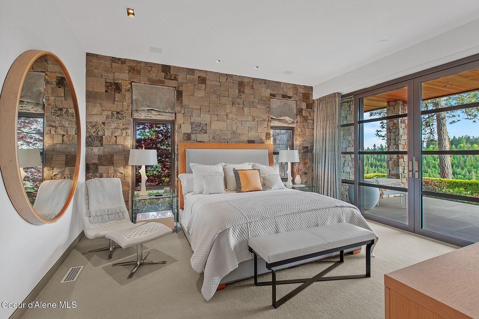 This is a primary bedroom featuring a stone accent wall, a large bed with white linens and an orange accent pillow, and a seating area with a modern chair and a round mirror. Large glass doors lead to an outdoor patio, offering natural light and views of the surrounding landscape. The room exudes a luxurious and serene atmosphere.