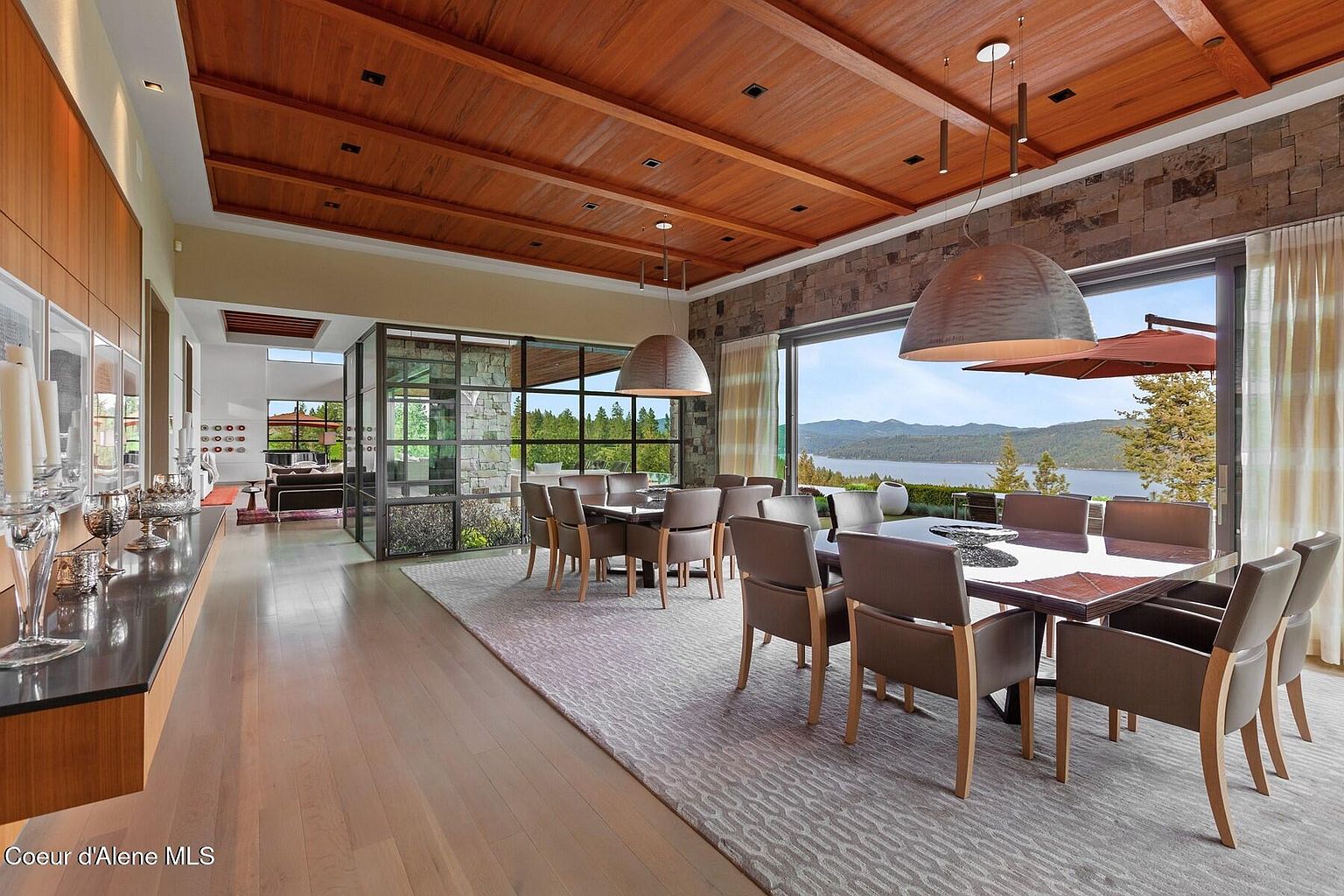 This is a spacious dining room featuring two large dining tables with seating for many people. The room has a modern design with a wood-paneled ceiling, stone accent wall, and large windows offering a scenic view of a lake and mountains. The room is well-lit by pendant lighting and natural light, creating an inviting atmosphere.