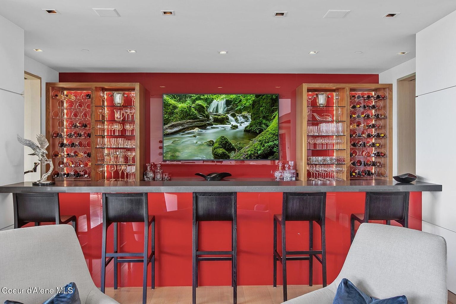 This image showcases a modern wine cellar or bar area featuring a striking red backsplash and a dark countertop. Wine racks and glass storage flank a large television displaying a nature scene, creating a focal point. Bar stools line the counter, and comfortable armchairs are positioned in the foreground, suggesting a sophisticated and inviting space for entertaining.