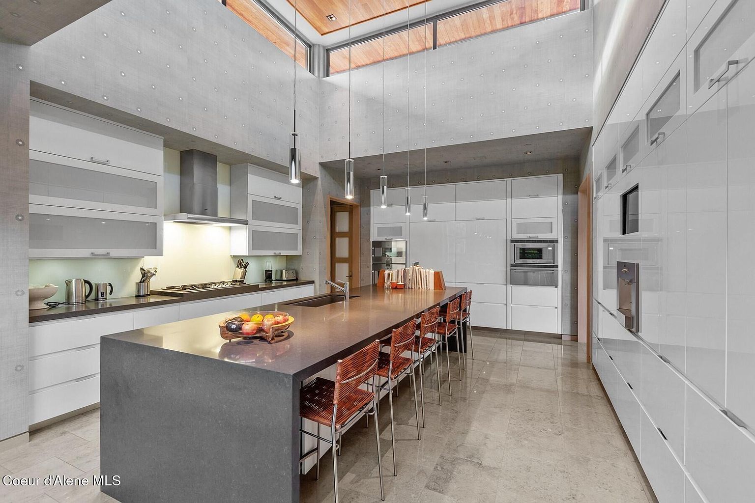 This is a modern kitchen with sleek, white cabinetry and a large gray island with seating. The kitchen features stainless steel appliances and unique pendant lighting. The high ceilings and large windows create a bright and airy space, emphasizing the contemporary design.