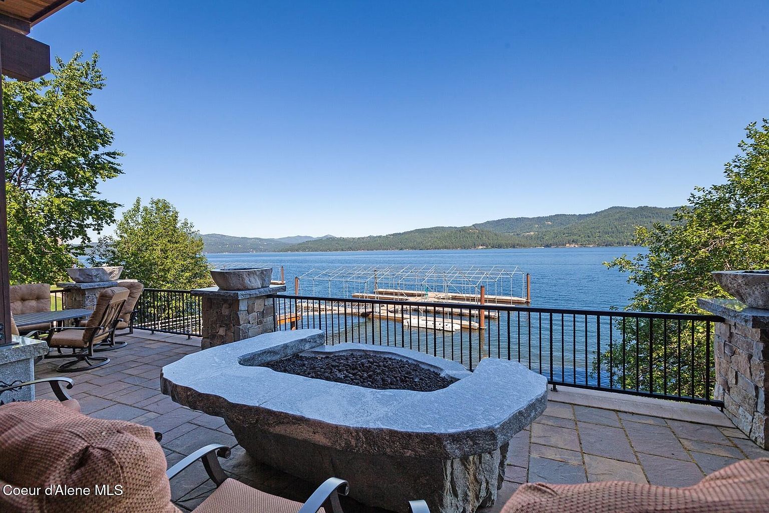 This image showcases a luxurious patio or deck area with a stunning lake view. A stone fire pit takes center stage, surrounded by comfortable seating. The patio features stone flooring and a black metal railing, enhancing the outdoor living space and providing a picturesque setting.