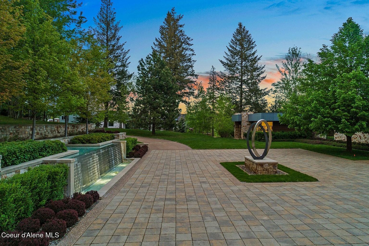 This image showcases a beautifully landscaped yard with a modern aesthetic. A striking water feature and a contemporary sculpture serve as focal points, complemented by lush greenery and mature trees. The paved driveway and manicured lawn create an inviting and luxurious outdoor space.