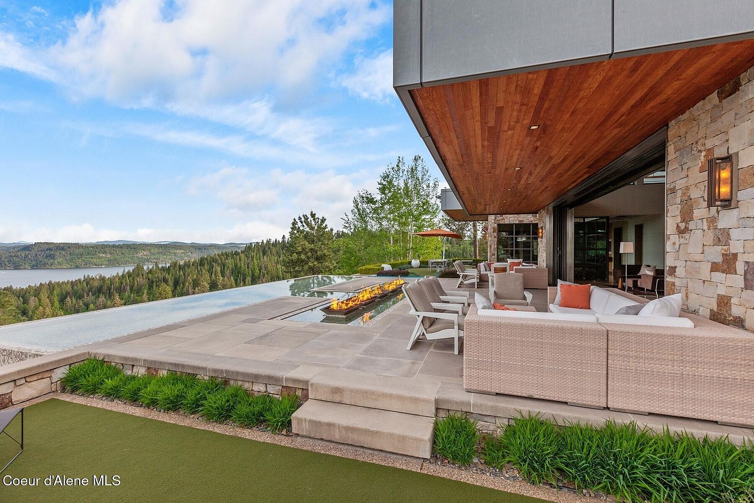 This image showcases a luxurious outdoor patio area with a stunning view of a lake and forest. The patio features modern furniture, a fire pit, and an infinity pool that blends seamlessly with the landscape. The architectural design incorporates natural stone and wood elements, creating a sophisticated and inviting space for relaxation and entertainment.