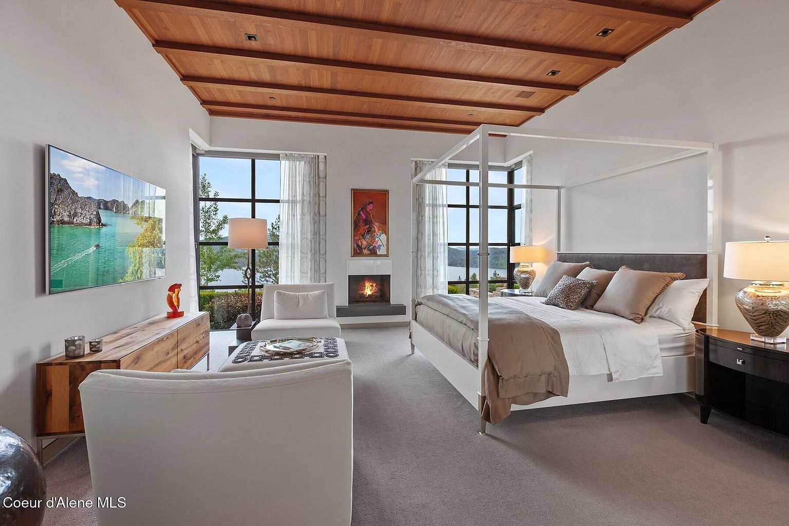 This is a luxurious primary bedroom featuring a four-poster bed, a modern fireplace, and large windows with scenic views. The room is decorated in neutral tones with wood accents on the ceiling, creating a serene and elegant atmosphere. A large TV is mounted on the wall, and comfortable seating is arranged near the fireplace.