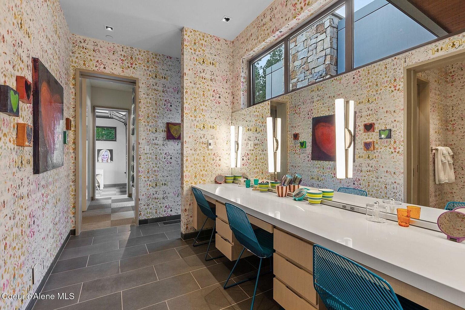 This is a well-lit primary bathroom featuring a long vanity with multiple seating areas and mirrors, complemented by unique wallpaper. The room has a modern design with clean lines and a playful use of color and pattern. The flooring is a dark tile, adding a touch of sophistication to the space.