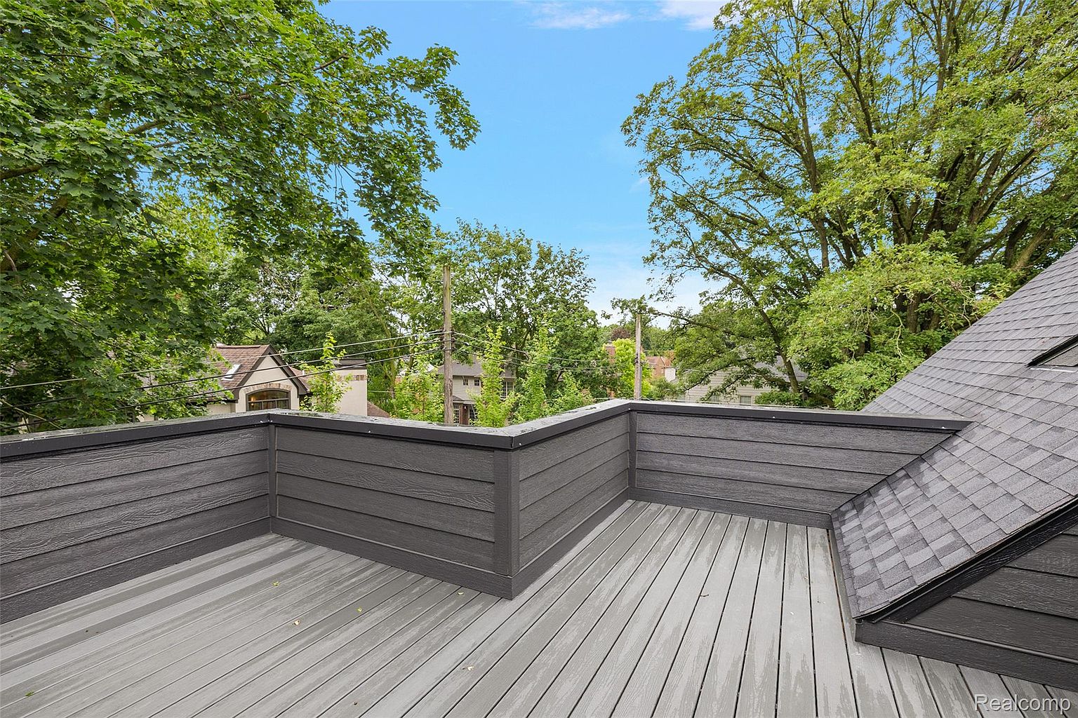 This image showcases a modern rooftop deck with gray composite decking and matching gray railings. The deck offers a view of the surrounding neighborhood and lush green trees under a bright blue sky. A section of the roof is also visible, adding to the architectural interest.