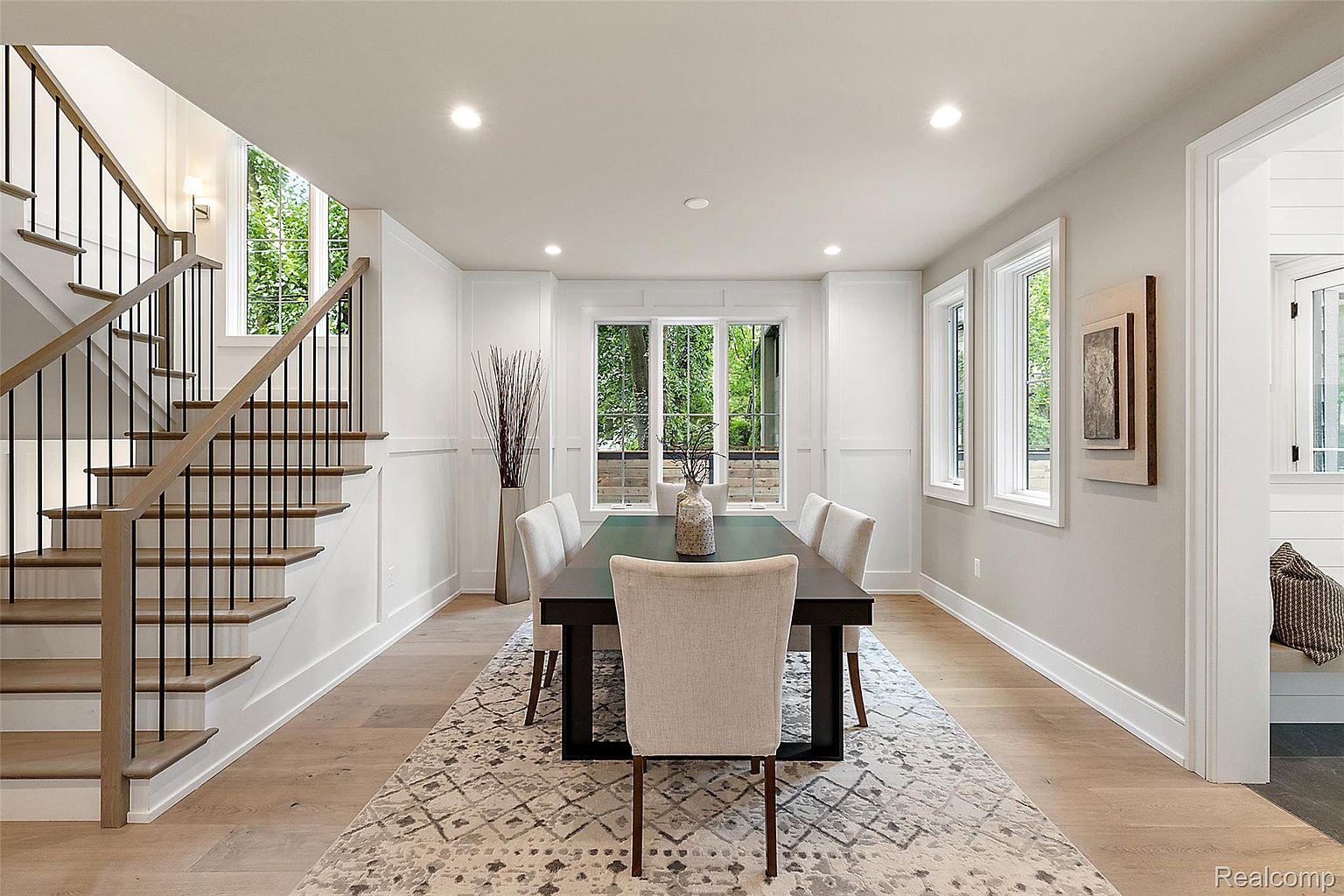 This is an interior shot of a dining room featuring a dark wood dining table surrounded by upholstered chairs, set on a patterned rug. Natural light floods the room through a large window and smaller adjacent windows, complemented by recessed lighting. A staircase is visible to the left, and an open doorway leads to another room, creating an inviting and spacious atmosphere.