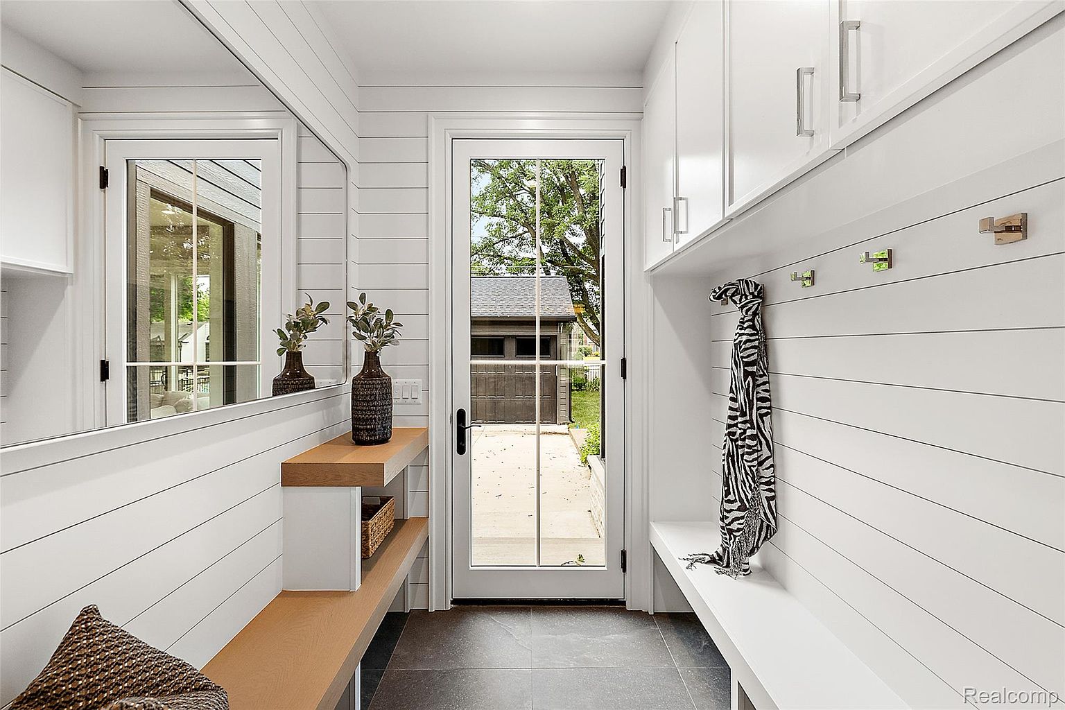 This interior shot showcases a modern laundry room with a clean and organized aesthetic. The room features white shiplap walls, built-in cabinetry, a wooden bench, and a mirror, creating a bright and functional space. A glass door provides natural light and a view to the outside, enhancing the room's appeal for potential buyers.