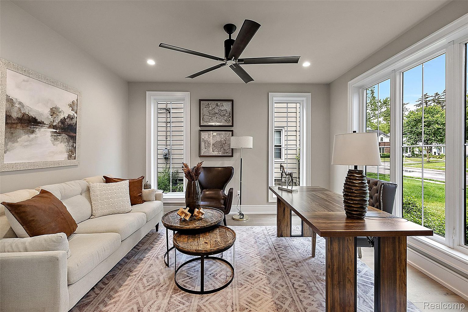This is an interior shot of a well-lit home office or study. The room features a neutral color palette, a large wooden desk, a comfortable sofa, and a round nesting coffee table. Large windows provide ample natural light and a view of the outdoors, creating a bright and inviting workspace.