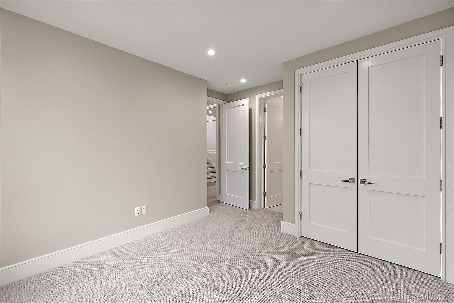 This is a well-lit bedroom featuring neutral-toned walls and light gray carpeting. The room includes a double door closet and an additional door leading to another room or hallway, suggesting ample storage and connectivity. The overall impression is clean, simple, and ready for personalization.