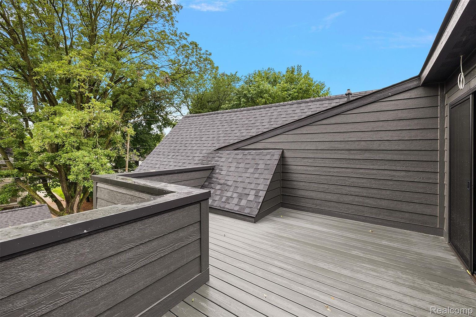 This image showcases a modern deck or balcony with gray wood planks and a matching gray railing. The deck is adjacent to a building with dark gray horizontal siding and a gray shingled roof. The view includes lush green trees in the background, creating a serene and private outdoor space.