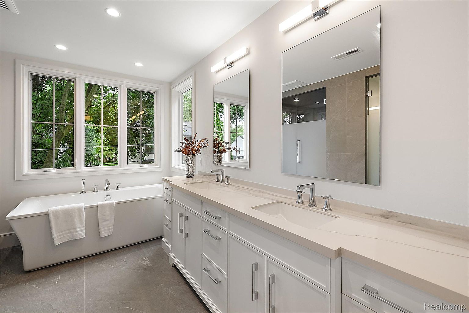 This is a bright and luxurious primary bathroom featuring a double vanity with white cabinetry and a light-colored countertop. Two large mirrors hang above the sinks, illuminated by modern light fixtures. A freestanding bathtub sits beneath a large window offering a view of lush greenery, creating a spa-like atmosphere.