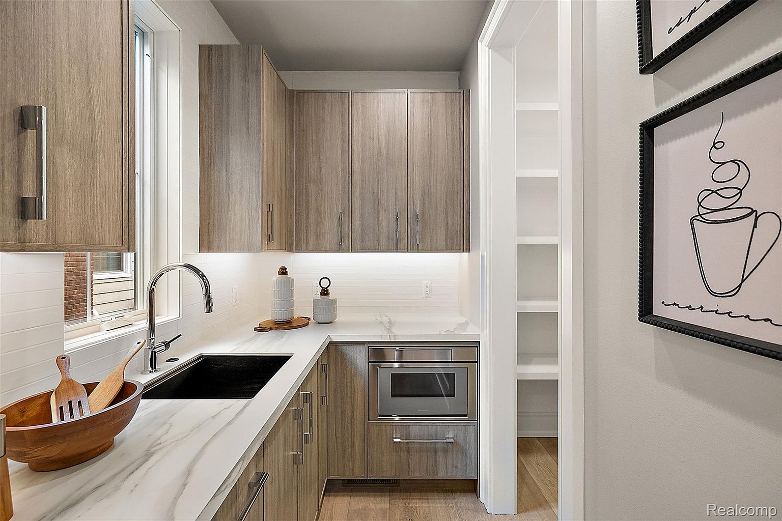 This is a well-lit kitchen featuring modern cabinetry with sleek, silver hardware and a light-colored countertop with a marble pattern. A stainless steel microwave is built into the lower cabinets, and a pantry is visible through an open doorway. The overall impression is clean, contemporary, and functional.