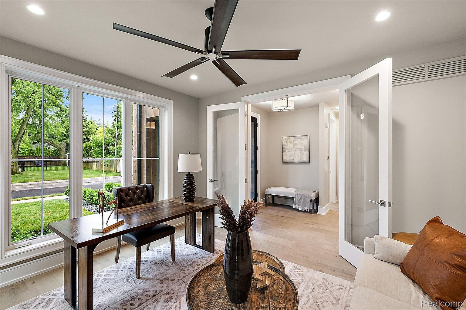This is an interior shot of a modern home office featuring a dark wood desk and chair, a patterned rug, and a round coffee table with decorative items. Large windows provide natural light and a view of the outdoors, while a doorway leads to another room, showcasing a bench and artwork. The room exudes a sophisticated and comfortable atmosphere, ideal for a productive workspace.
