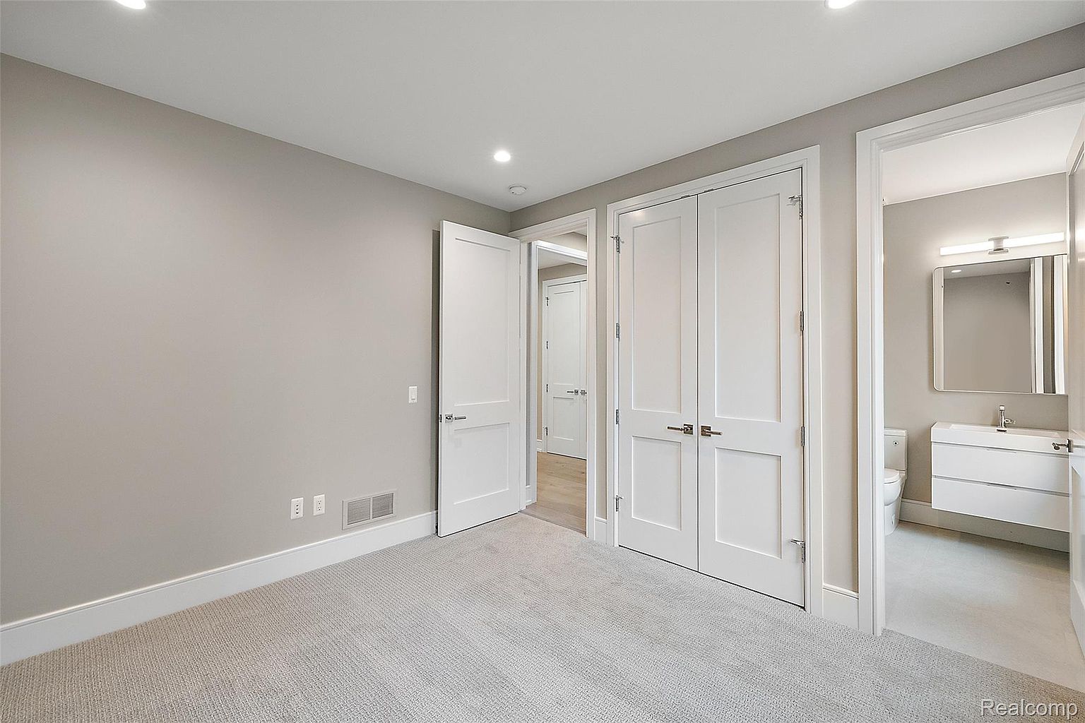 This is an interior shot of a bedroom featuring neutral gray walls and light gray carpet. The room has white trim and baseboards, with white doors leading to a closet and a bathroom. The overall impression is clean and modern, creating a serene and inviting space.