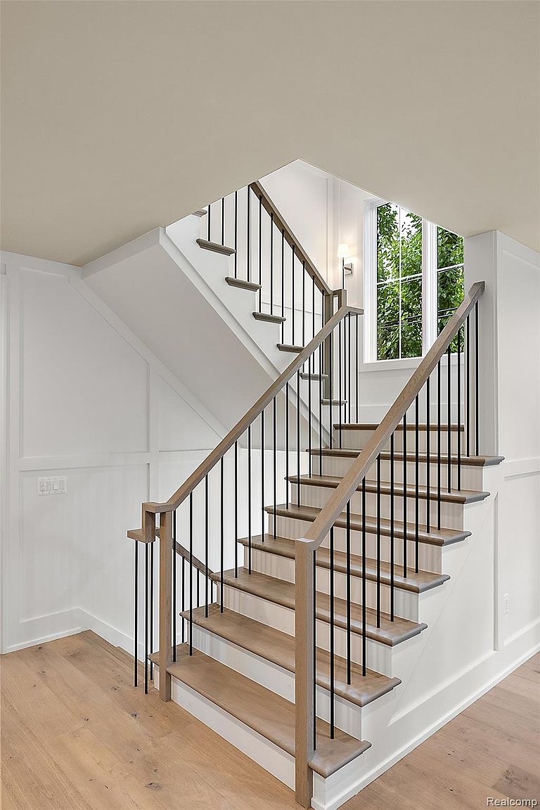 This interior shot showcases a well-lit staircase with light wood treads and white risers, complemented by a dark metal railing and light wood handrail. The walls are painted white with wainscoting detail, and natural light streams in from a nearby window, creating a bright and inviting atmosphere. The hardwood flooring on the landing adds warmth and continuity to the design.