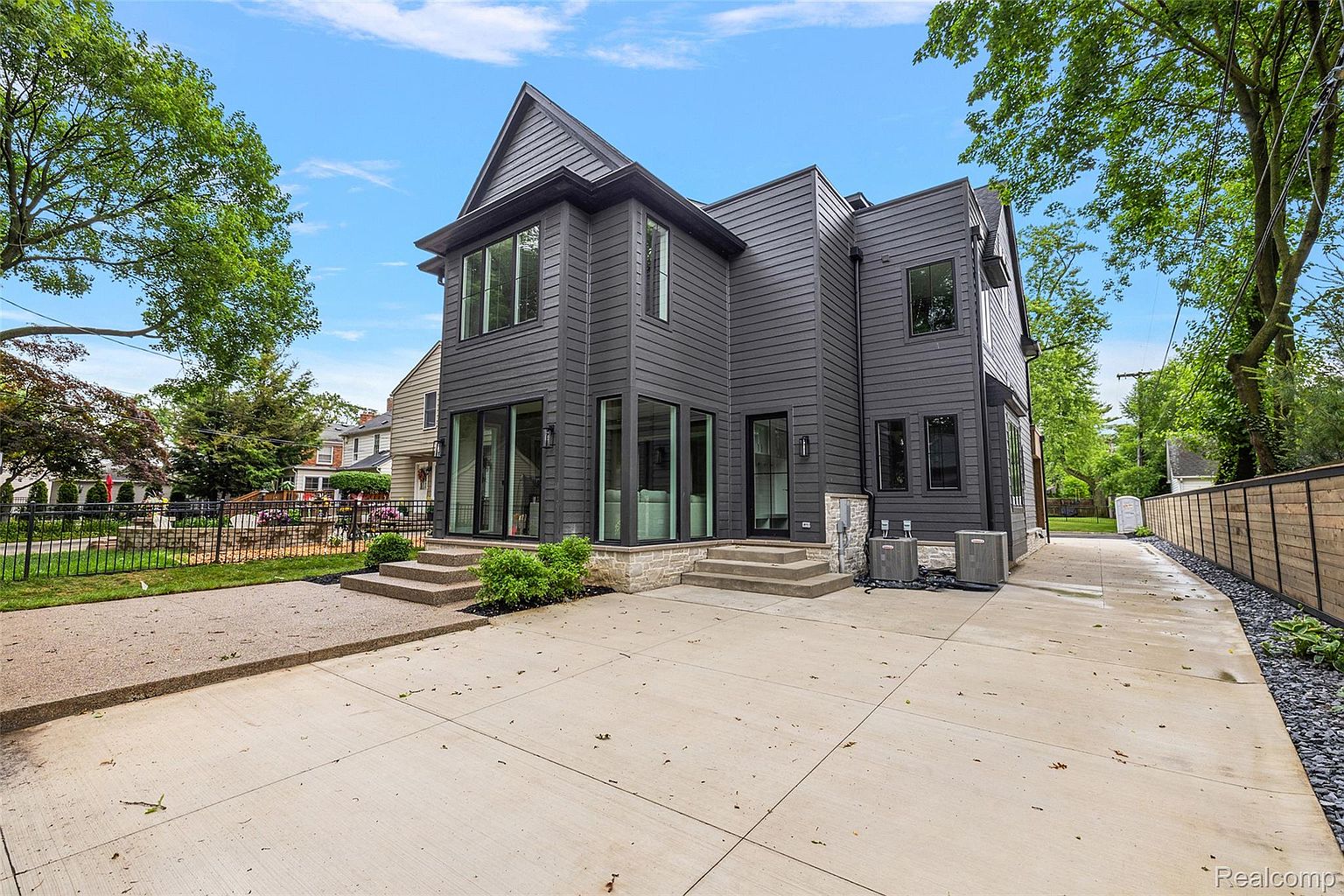 This is a rear view of a modern, two-story house with dark gray siding and black-framed windows. The property features a concrete patio area with steps leading up to the house, and a well-maintained lawn and landscaping. The exterior design is sleek and contemporary, offering a sophisticated and inviting appearance.