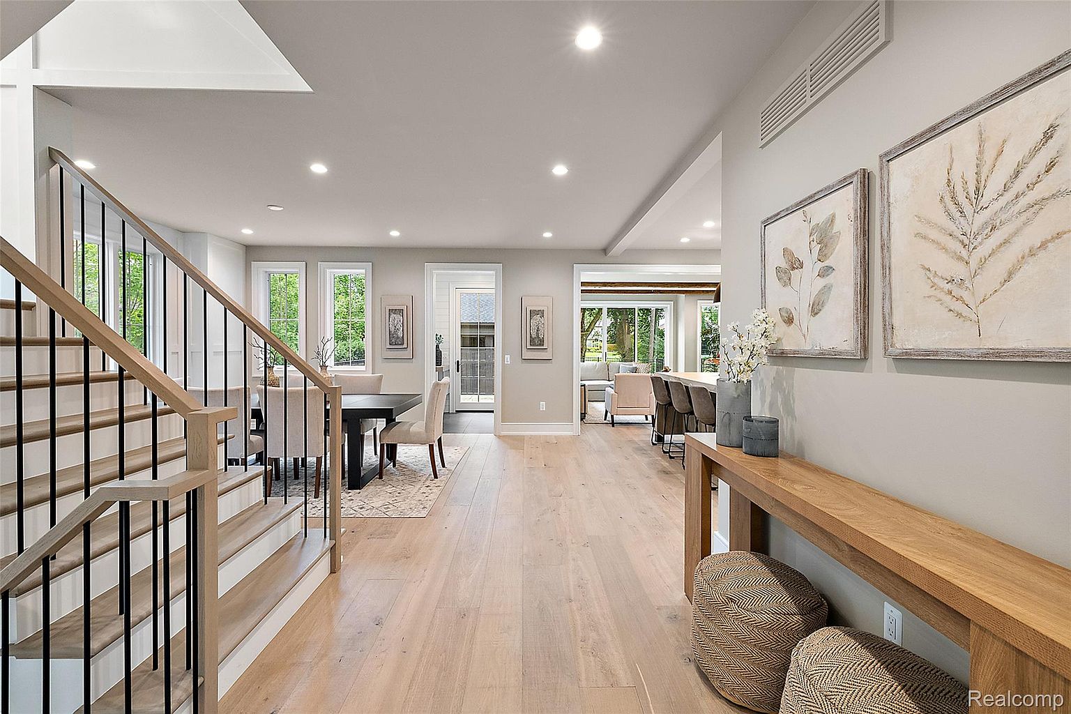 This interior shot showcases a bright and open hallway with a staircase on the left. The hallway features light wood flooring, neutral-toned walls, and artwork displayed on the right. The staircase has light wood steps and black metal railings, adding a modern touch to the space.