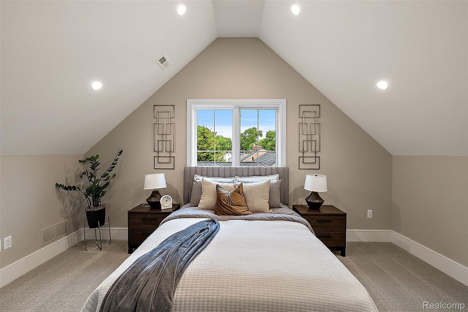 This is a cozy bedroom featuring a bed with a gray headboard, white quilted bedding, and a gray throw blanket. Two wooden nightstands with lamps flank the bed, and decorative metal wall art pieces hang on either side of the window. The room has a neutral color palette and an attic-style ceiling, creating a warm and inviting atmosphere.