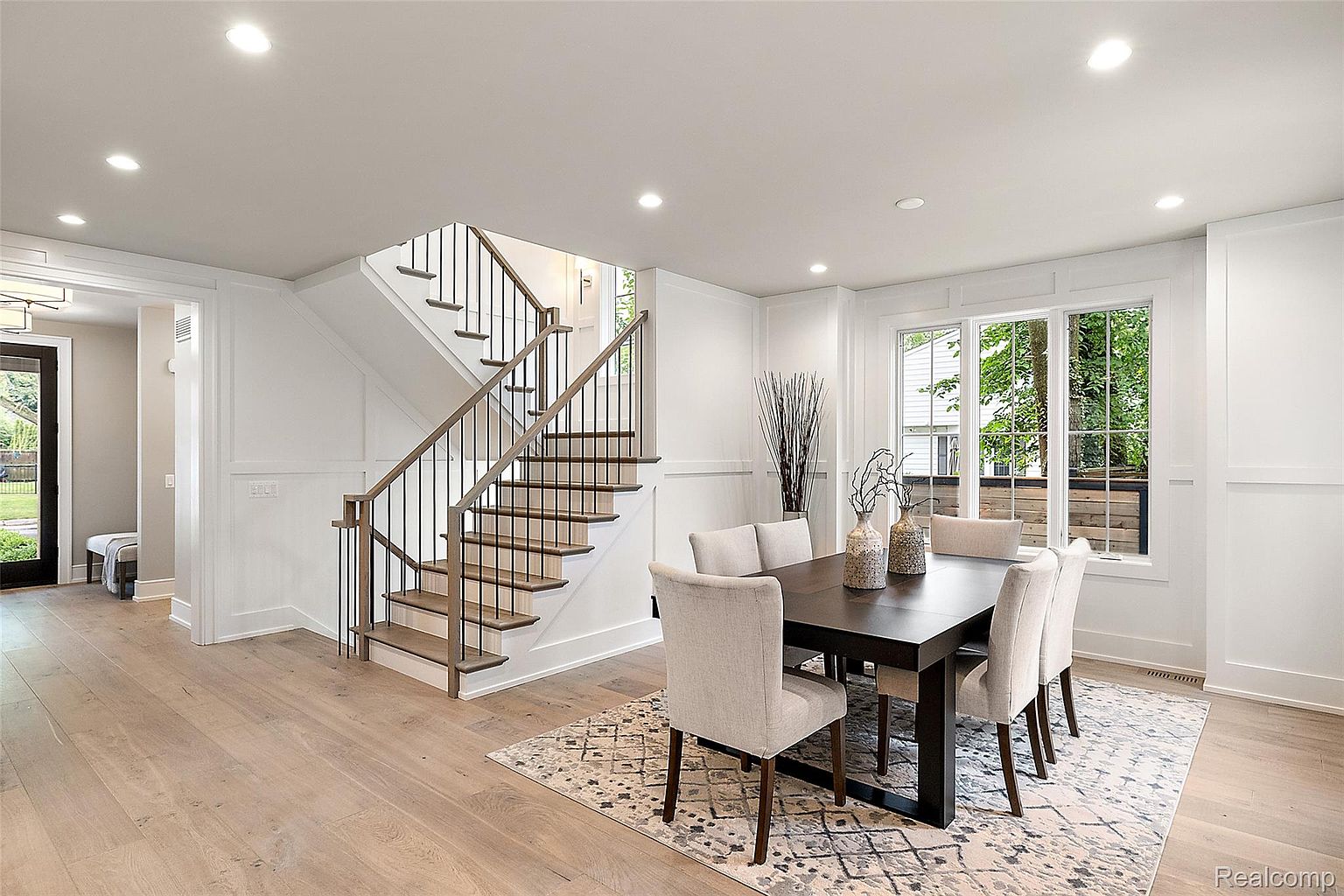 This is an interior shot of a dining room featuring a dark wood dining table with six upholstered chairs, set on a patterned area rug. A staircase is visible to the left, and a large window provides natural light. The room is decorated in a neutral color palette, creating a bright and inviting space.