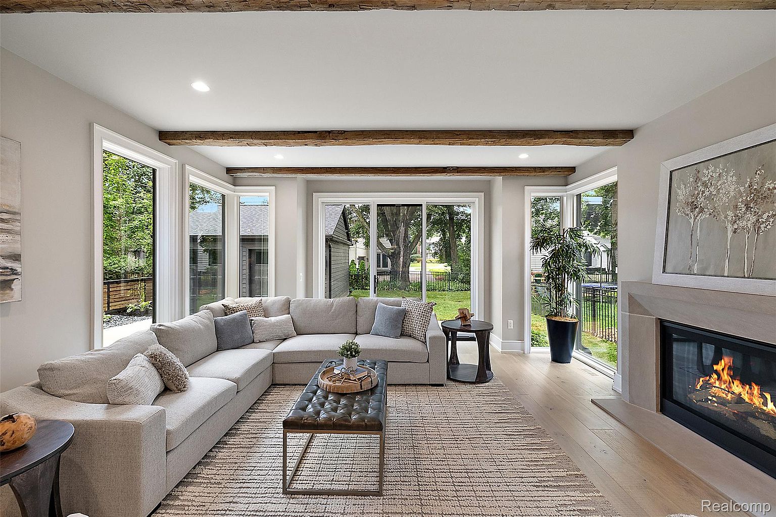 This is a bright and inviting living room featuring a large sectional sofa, a tufted ottoman coffee table, and a neutral-toned area rug. The room is well-lit with natural light streaming through large windows that offer views of the outdoors. Exposed wooden beams add a touch of rustic charm to the space, which also includes a modern fireplace.