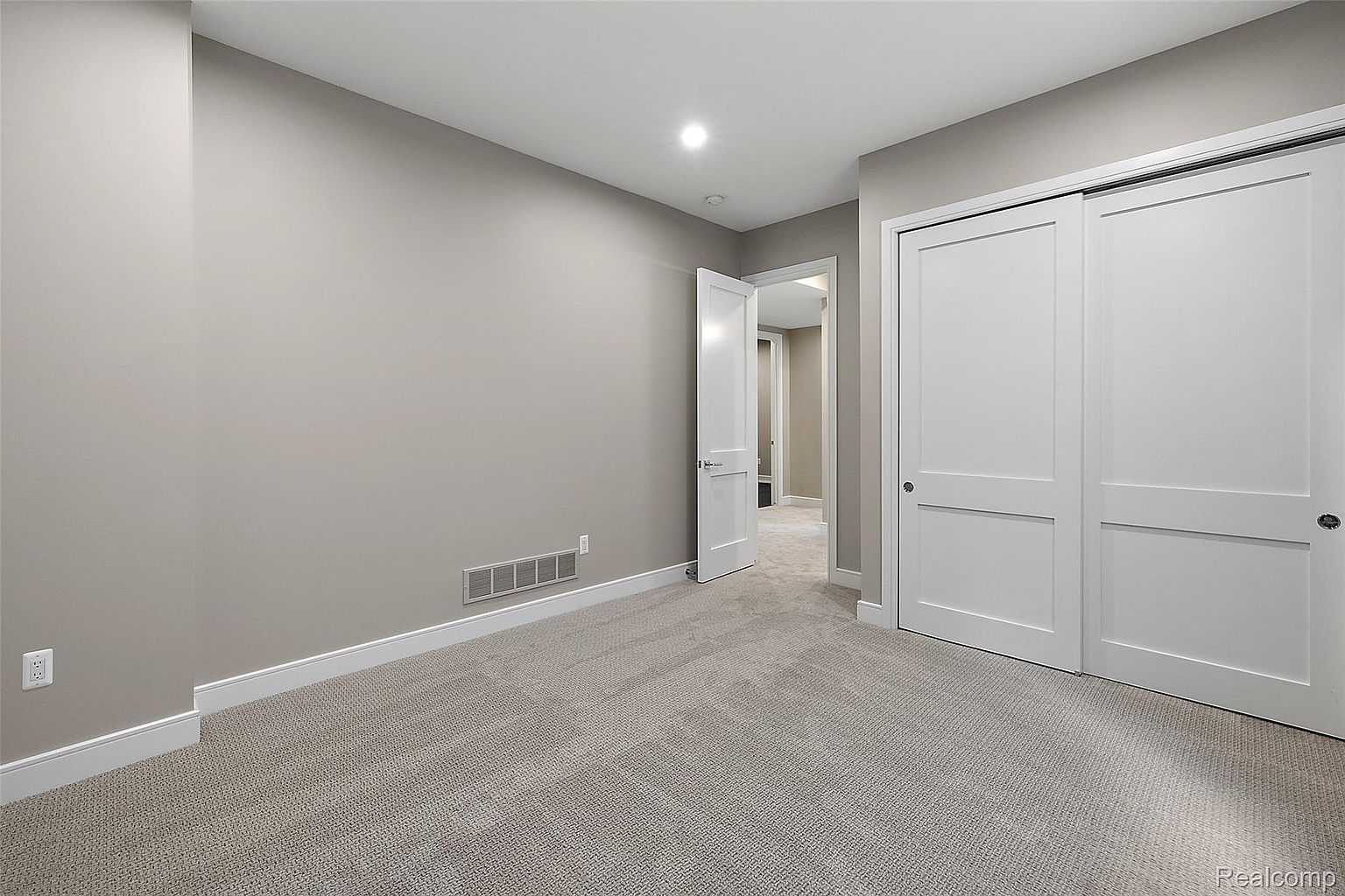 The image showcases a well-lit bedroom with neutral-toned walls and carpet. A white door is slightly ajar, leading to another room, while a large closet with sliding white doors occupies the right side of the frame. The room appears clean and spacious, suitable for a guest or child's bedroom.