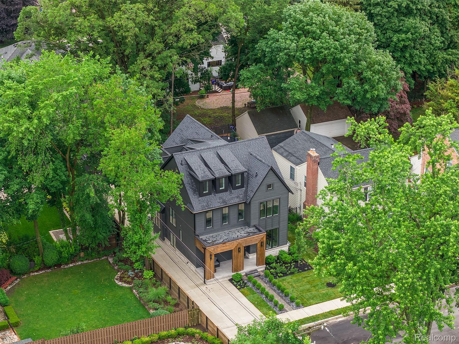 This aerial view showcases a modern two-story home with a dark gray exterior, complemented by a gray roof with dormer windows. A wooden portico covers the entryway, and a well-manicured lawn and landscaping surround the property. The long driveway leads to the street, and mature trees provide privacy and shade.