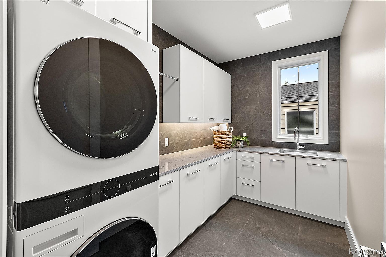 This is a modern laundry room featuring a stacked washer and dryer unit, sleek white cabinetry with minimalist hardware, and a gray countertop. The room is well-lit with a combination of natural light from a window and under-cabinet lighting, complemented by dark gray tiled walls and flooring, creating a clean and functional space.