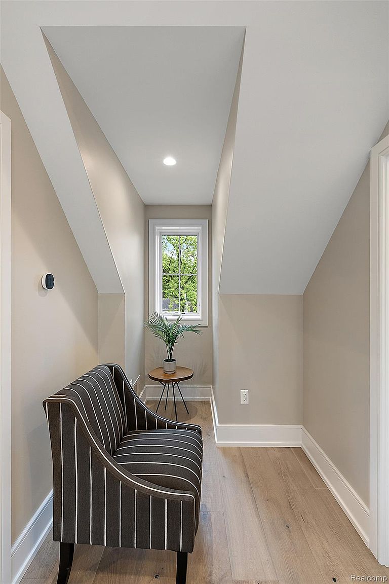 This interior shot showcases a cozy hallway nook featuring a striped armchair, a small round table with a potted plant, and a window providing natural light. The walls are painted in a neutral tone, complemented by white trim and light hardwood flooring. The space is well-lit and appears to be a comfortable and inviting area within the home.